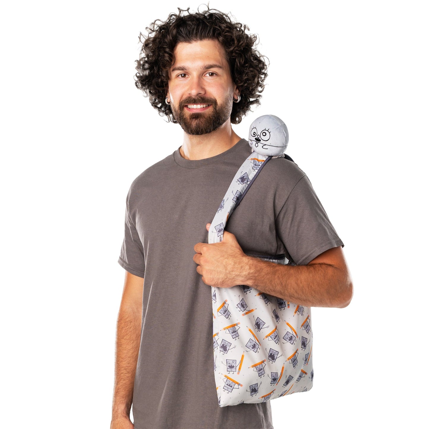 A man with curly hair in a gray shirt carries a Plushiverse DoodleBob Plushie Tote Bag from Nickelodeon, made from recycled plastic bottles, with a TeeTurtle plushie attached to the strap.