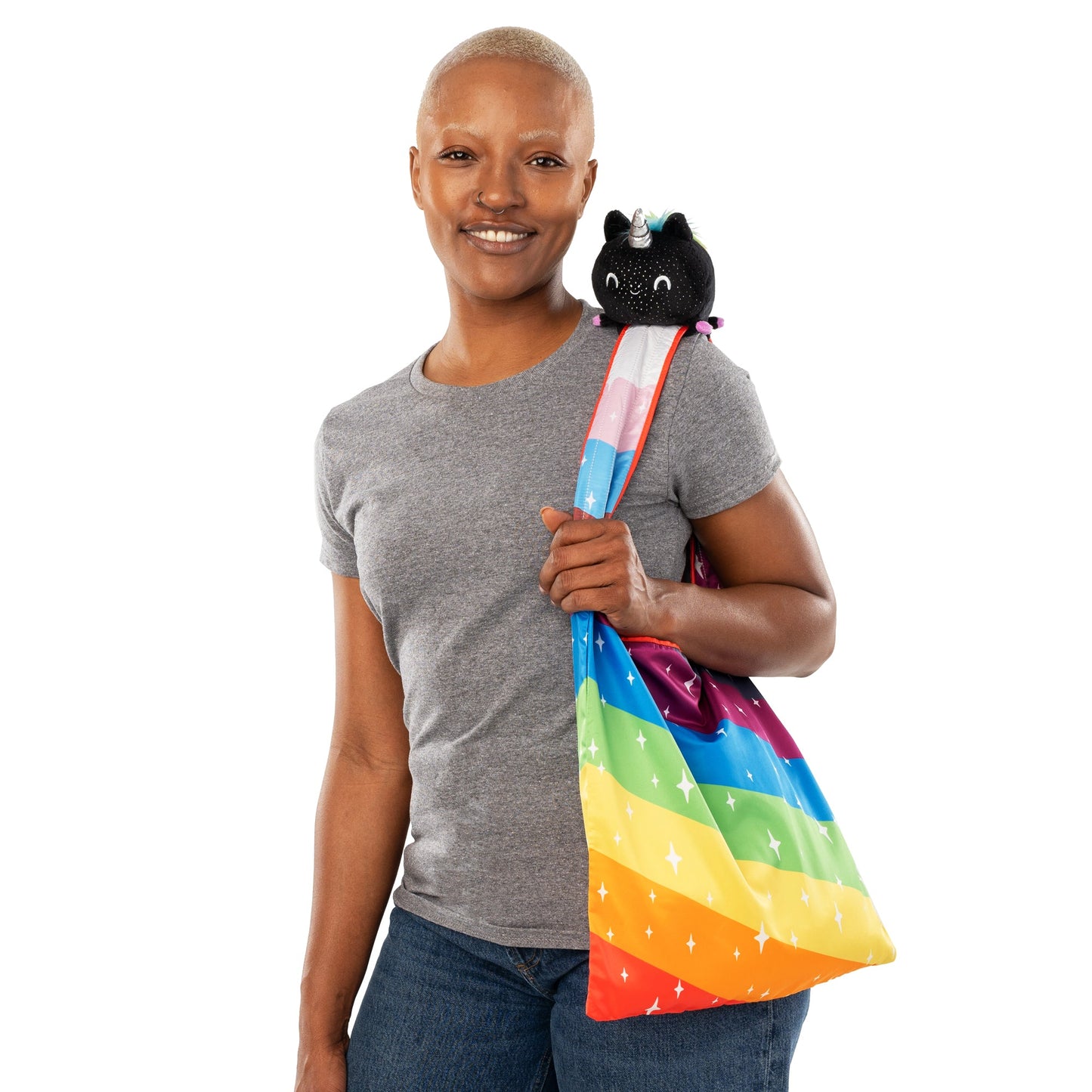 Person smiling, holding a rainbow-colored Plushiverse Unicorn Pride Plushie Tote Bag with a small black TeeTurtle plushie peeking out from the top over their shoulder.