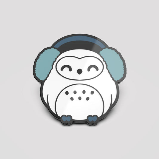 A white owl with blue TeeTurtle earphones on a gray background and Happy Hoo-lidays enamel pins.