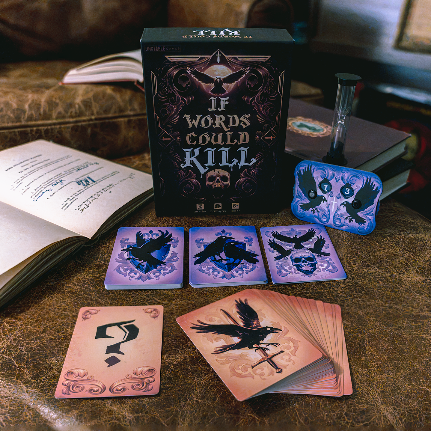 A game box labeled 'If Words Could Kill,' with a score tracker, hourglass and several cards spread out in front, in front of an open book, and a small bottle on a brown leather couch in an indoor setting.