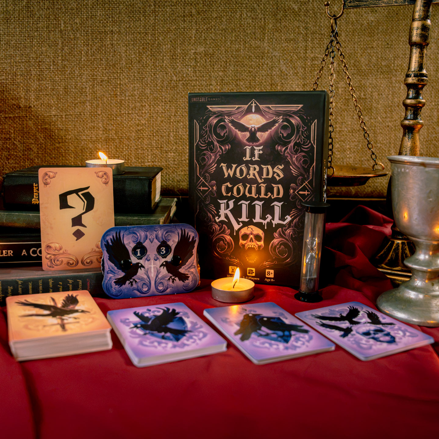 A tabletop scene showing a game box labeled 'If Words Could Kill,' with a score tracker, hourglass and several cards spread out in front, displayed on a red draped table against a burlap backdrop. There are lit candles, a stack of books and a scale of justice prop in the background. 