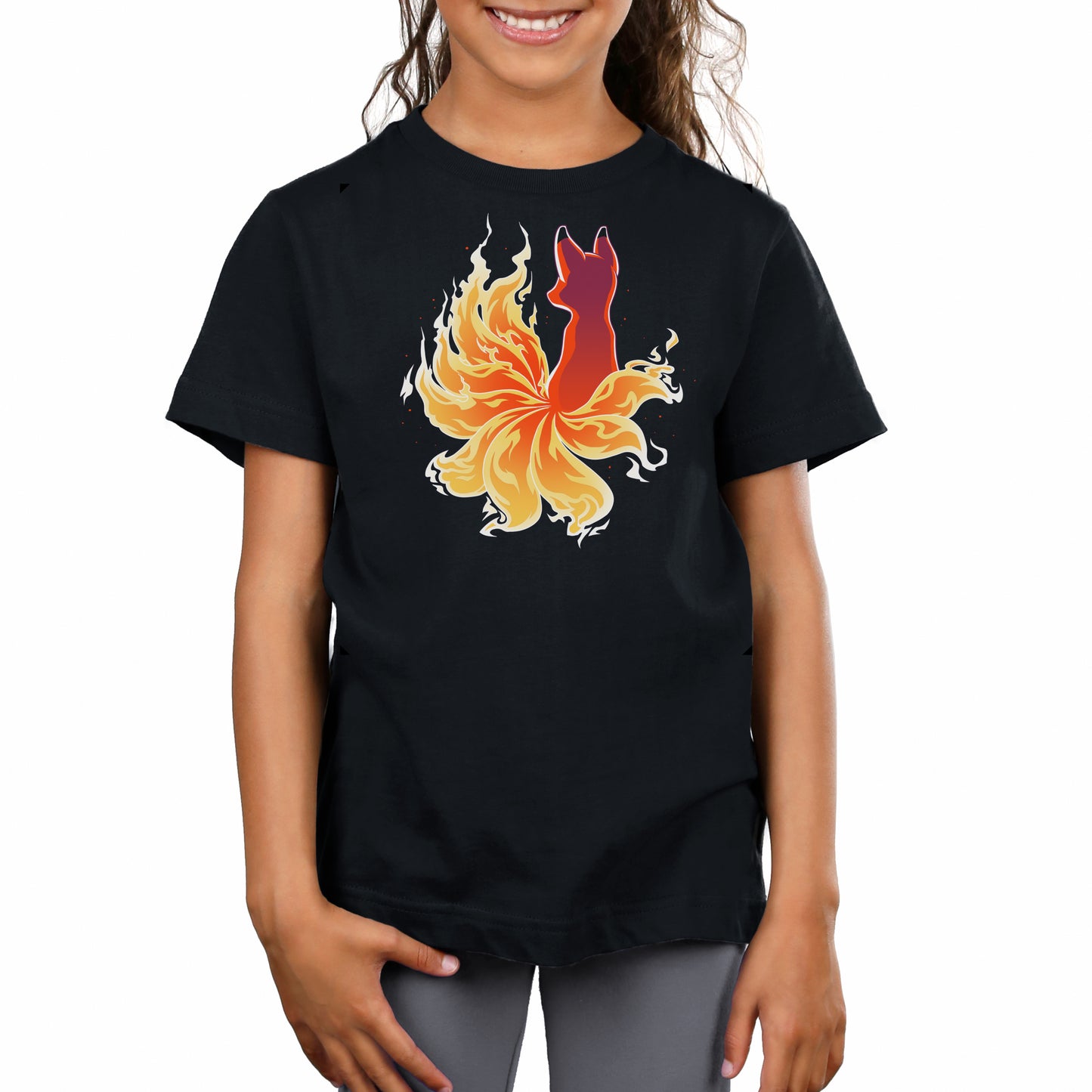 A child wearing a Fire Kitsune unisex tee by monsterdigital with a vibrant graphic of a red Fire Kitsune surrounded by yellow flames stands smiling, enjoying the super soft ringspun cotton fabric.