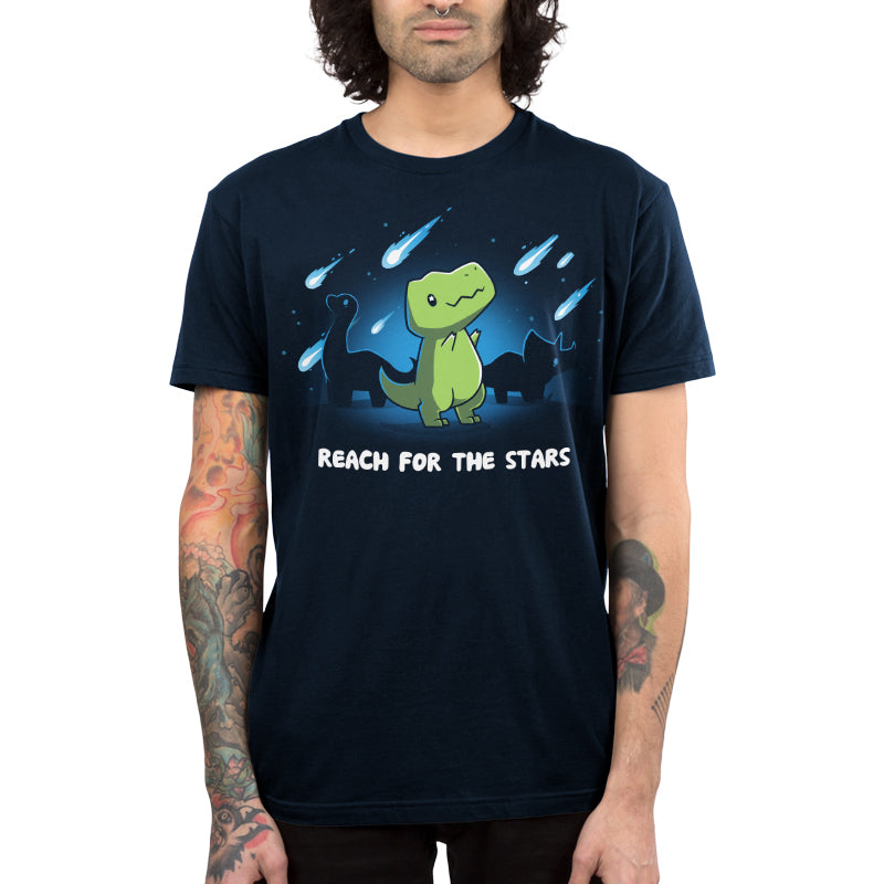 Person wearing a monsterdigital Reach For The Stars navy blue tee with a cartoon dinosaur and the text "Reach for the Stars". The super soft ringspun cotton shirt complements their tattoos and shoulder-length wavy hair perfectly.