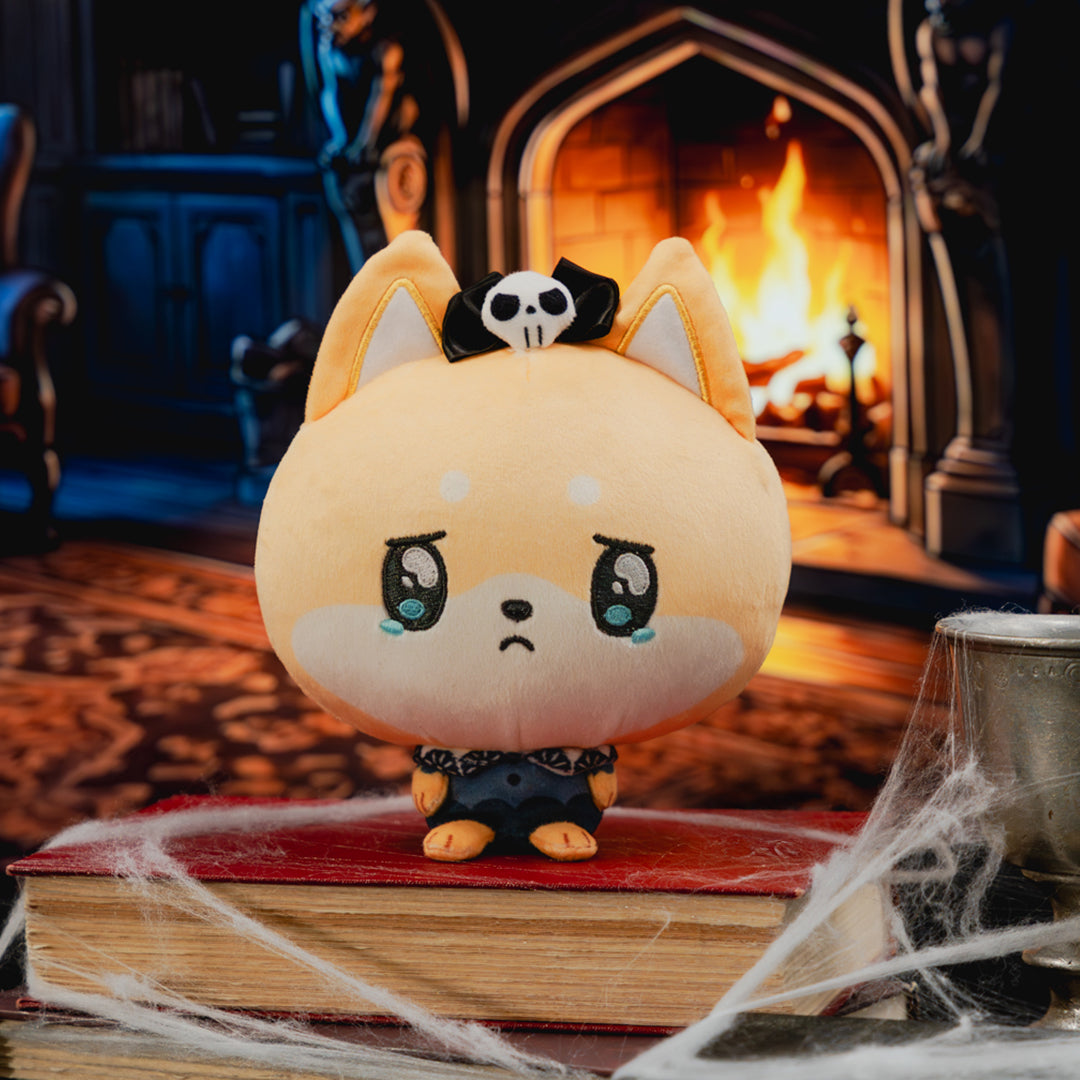 A cute, stylized plush toy with a round tan face, large dark eyes, and a small black-and-white skull on its head, posed on a red book with cobweb decor in front of a fireplace.