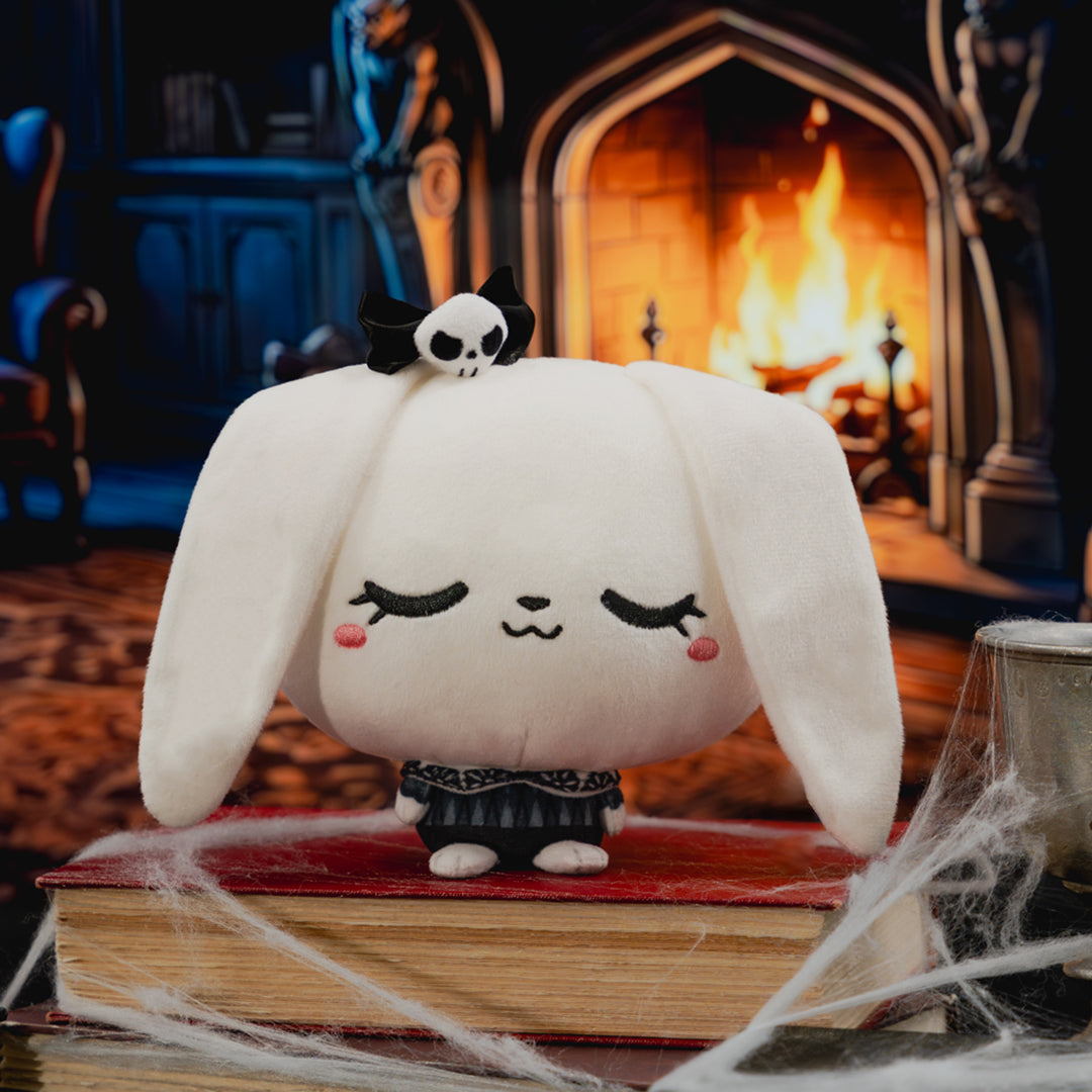 A white plush bunny character with long floppy ears, closed embroidered eyes, pink cheeks, and a small black bow on top, standing on a red book in a cozy fireplace scene with cobwebs in the foreground.