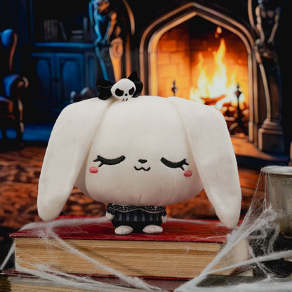 A white plush bunny character with long floppy ears, closed embroidered eyes, pink cheeks, and a small black bow on top, standing on a red book in a cozy fireplace scene with cobwebs in the foreground.