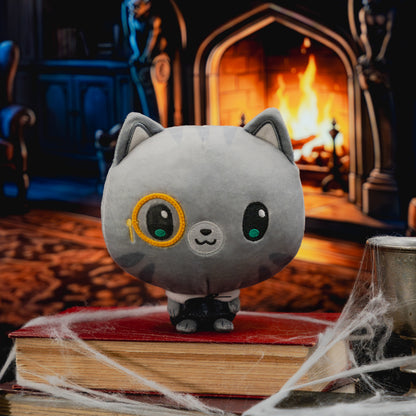 A gray cat plush toy with a single yellow-rimmed monocle and large glossy eyes, posed on top of stacked red books with cobwebbing in front, set against a dark cozy room with a burning fireplace in the background.