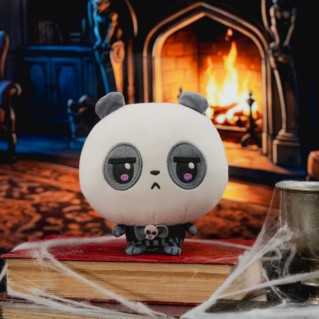 A cute, round white plush panda toy with sad, grumpy eyes and small gray ears sits on a red book surrounded by fake cobwebs, with a cozy, dark fireplace in the background.