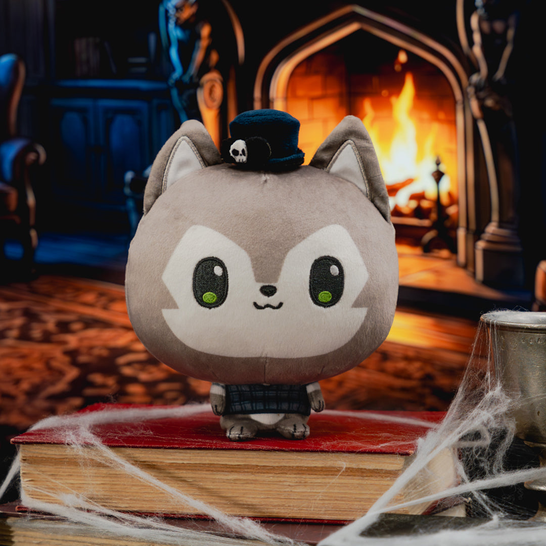 A cute, round gray plush toy with large white eyes, green highlights, and a small black top hat with a skull emblem, standing on a red book surrounded by fake cobwebs in a cozy, dark, gothic room with a burning fireplace in the background.