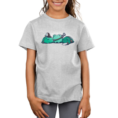 A young girl wearing a light gray kids T-shirt featuring a cartoon graphic of three green frogs lounging and carrying small items, called RPG Frogs by monsterdigital.