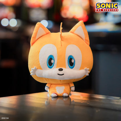 Tails Cloudhead, a plushie with a big head and a tiny body. He has a little smile on his face. He is sitting on a table with blurry arcade machines behind him. 