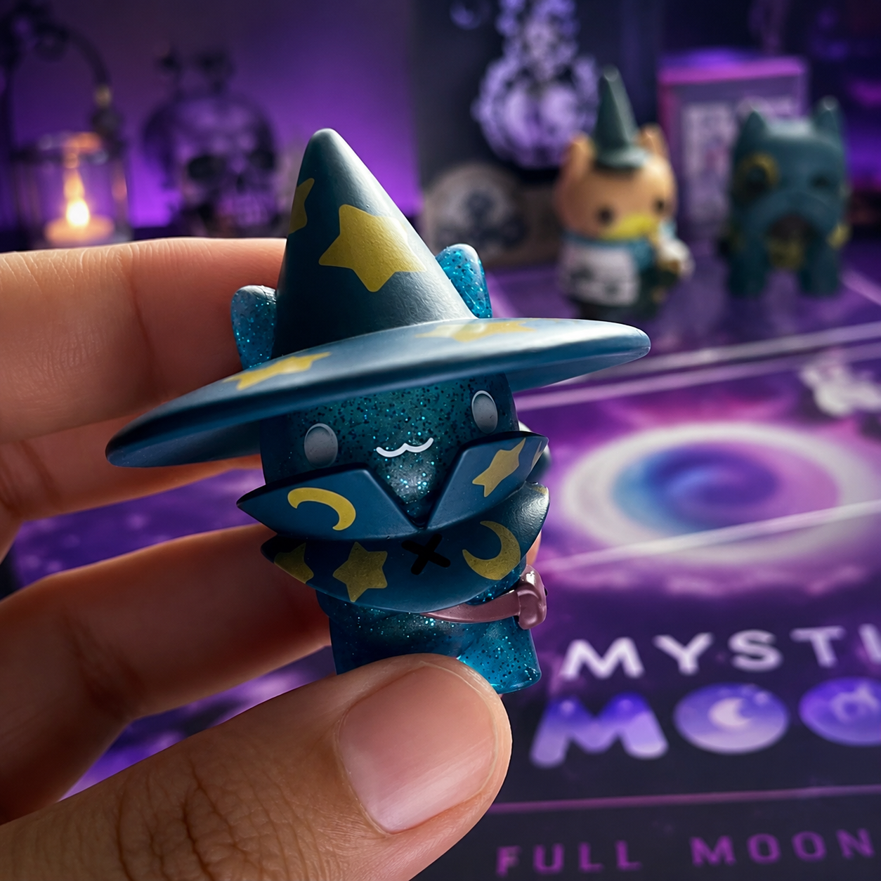 A hand holds a glittery Mystic Moon vinyl figure with a pointed hat decorated with yellow stars and crescent moons, set against a purple themed background with other collectible figures.