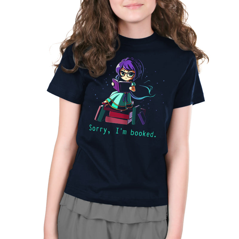 A person wearing a navy blue T-shirt made from super soft ringspun cotton, featuring an illustration of a character sitting on a pile of books and text that reads "Sorry I'm Booked" from monsterdigital.