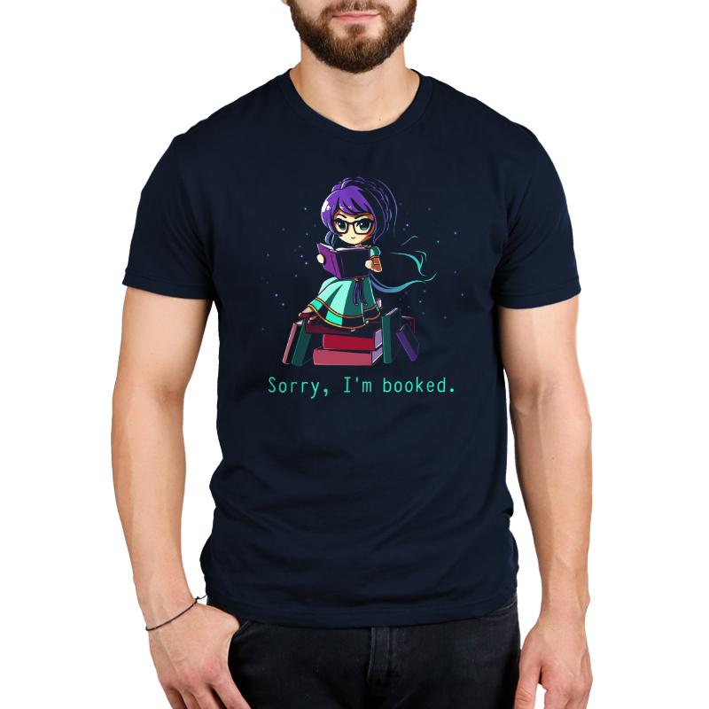 A bearded man wearing a super soft ringspun cotton navy blue T-shirt with an illustration of a character reading a book on top of a stack of books and the text "Sorry, I'm booked," by monsterdigital.