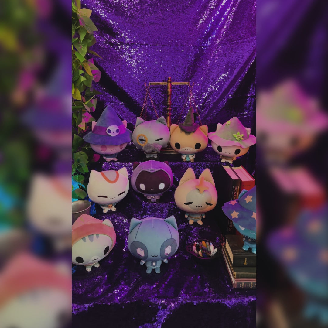 The whole set of 10 Mystic Moon Cloudheads plushies in front of a sparkly purple background. Also visible are books, a salt lamp, a crystal ball, an antique scale and a leafy green plant. 
