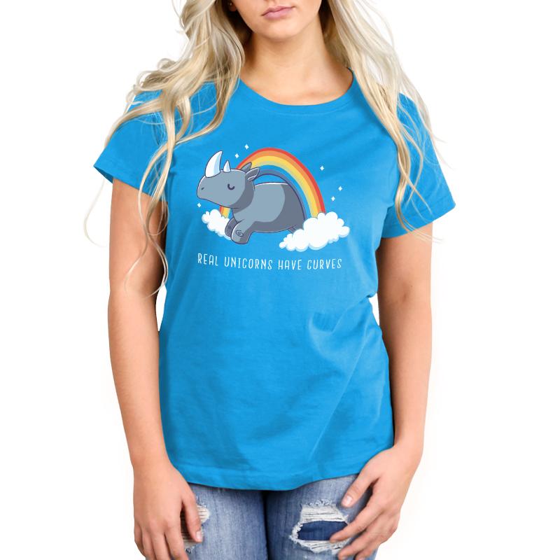 Woman wearing a cobalt blue Real Unicorns Have Curves t-shirt made from super soft ringspun cotton by monsterdigital, featuring a graphic of a rhinoceros, rainbow, and the text "Real Unicorns Have Curves." She is also wearing ripped jeans.
