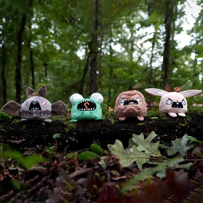 Twisted Cryptids: Mothman 4" Reversible Plushie, Twisted Cryptids: Loveland Frog 4" Reversible Plushie, Twisted Cryptids: Bigfoot 4" Reversible Plushie, and Twisted Cryptids: Jackalope 4" Reversible Plushie sitting on a mossy log in a forest